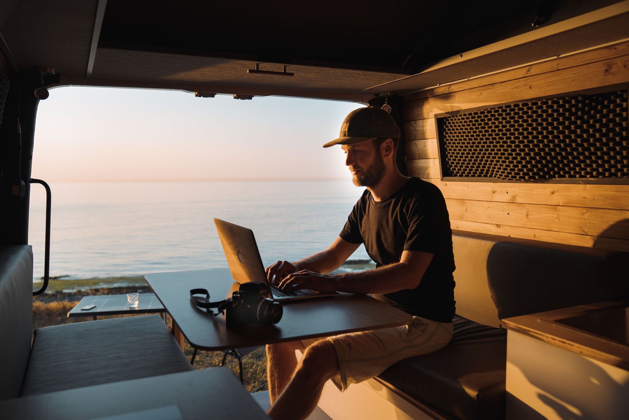 Mann arbeitet im energieautarken Wohnmobil mit Laptop und Blick auf das Meer bei Sonnenuntergang, betrieben durch Generatoren und Photovoltaik.