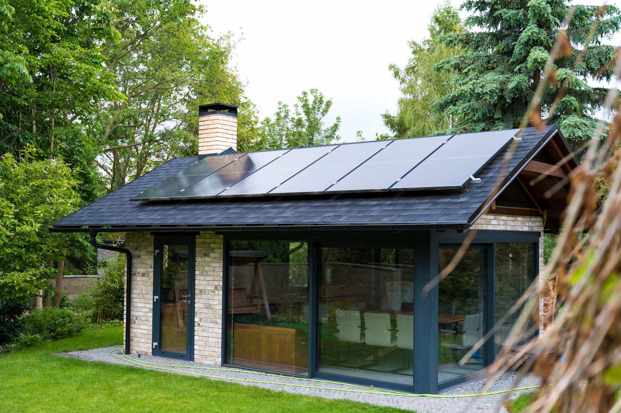 Kleines modernes Haus mit Photovoltaik-Anlagen auf dem Dach, umgeben von Bäumen und Garten.