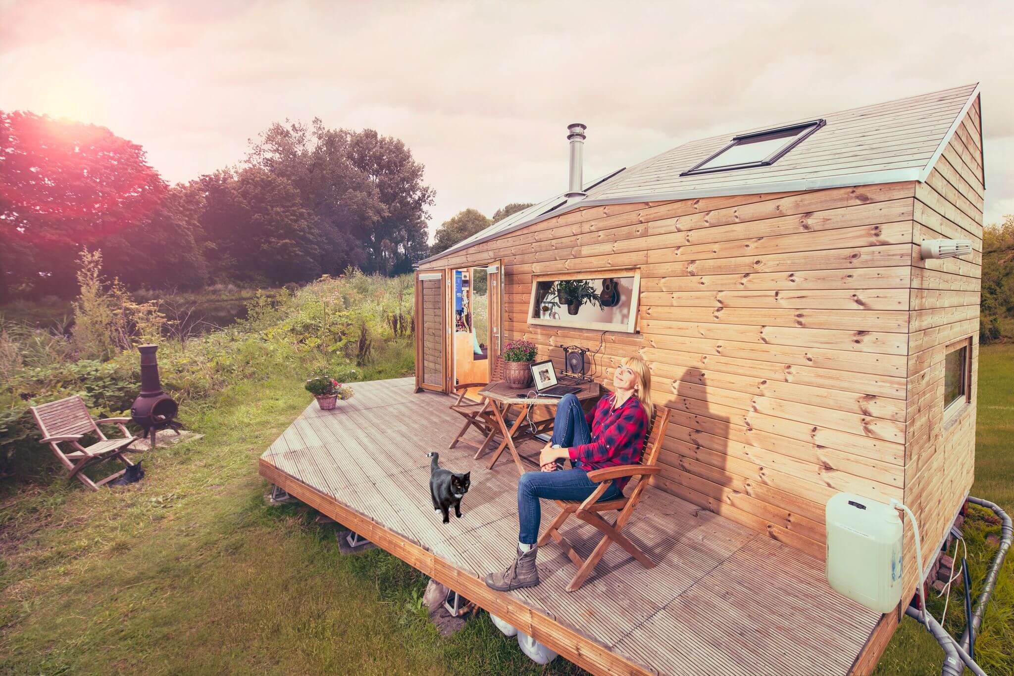 Eine Frau sitzt neben einem enegrieautarken Tiny House mit Holzverkleidung in ländlicher Umgebung.